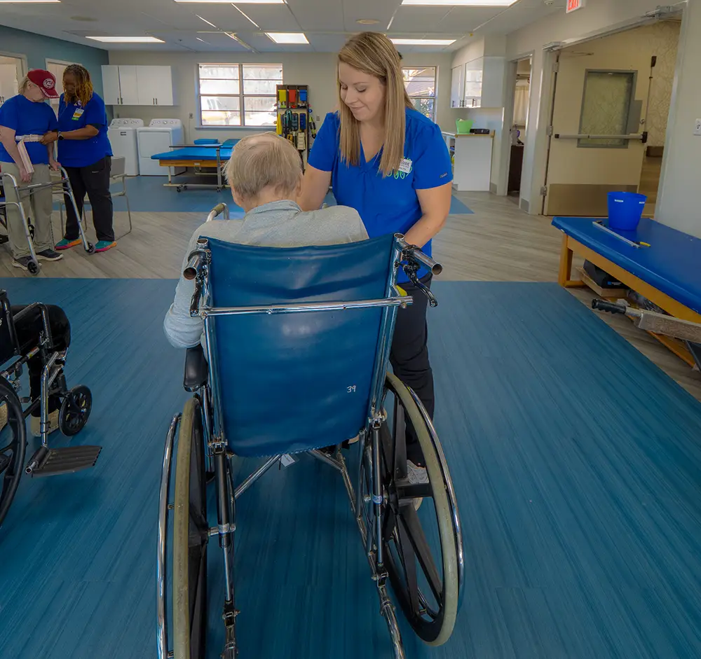A healthcare worker assists an older adult seated in a wheelchair in a rehabilitation facility with therapy tables and other staff in the background.