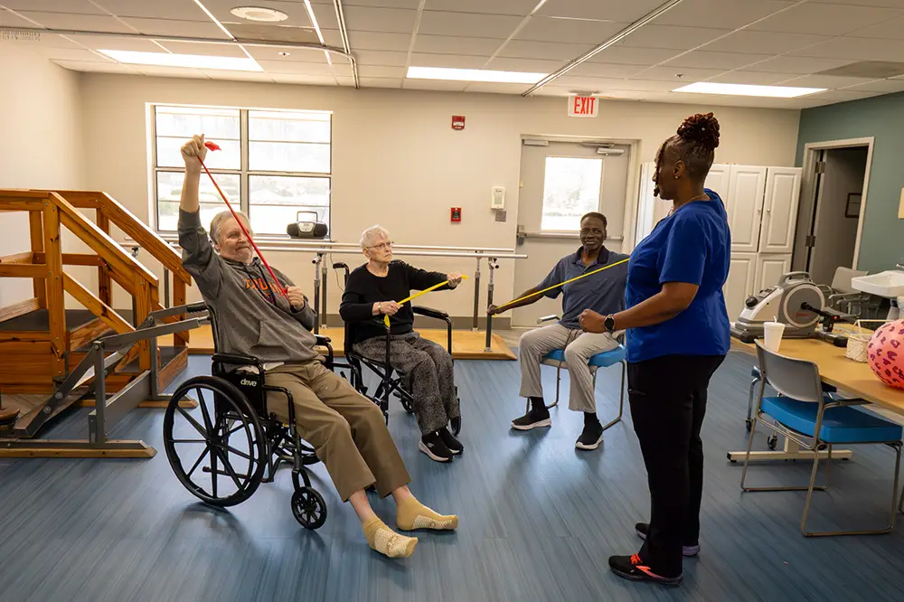 Three older adults, two in wheelchairs, exercise with resistance bands led by a female instructor in a brightly lit physical therapy room.