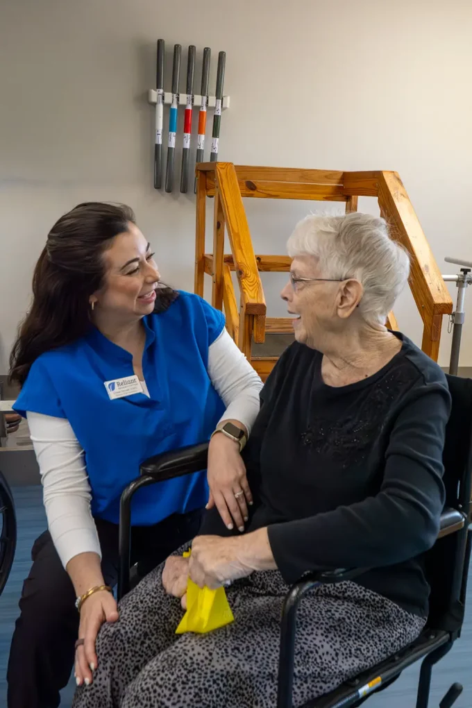 A healthcare worker in blue scrubs smiles and talks with an elderly woman in a wheelchair in a physical therapy room.