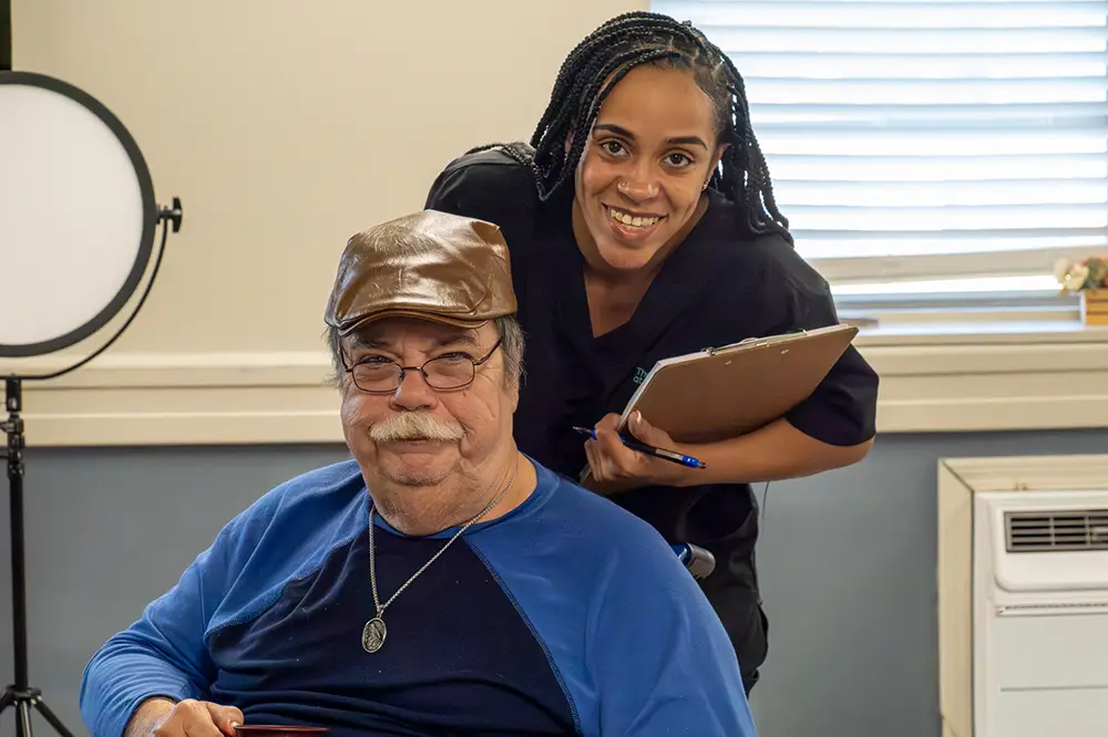 An older man in a blue shirt and cap sits in a wheelchair while a smiling woman in scrubs stands behind him, holding a clipboard.