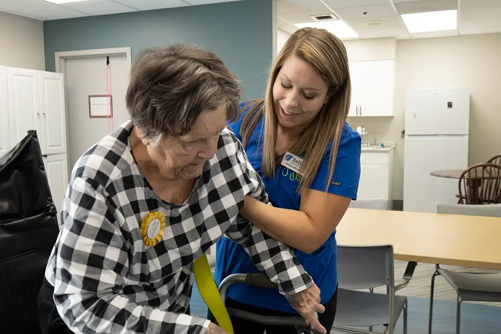 A caregiver in blue scrubs assists an elderly woman using a walker in a healthcare facility.
