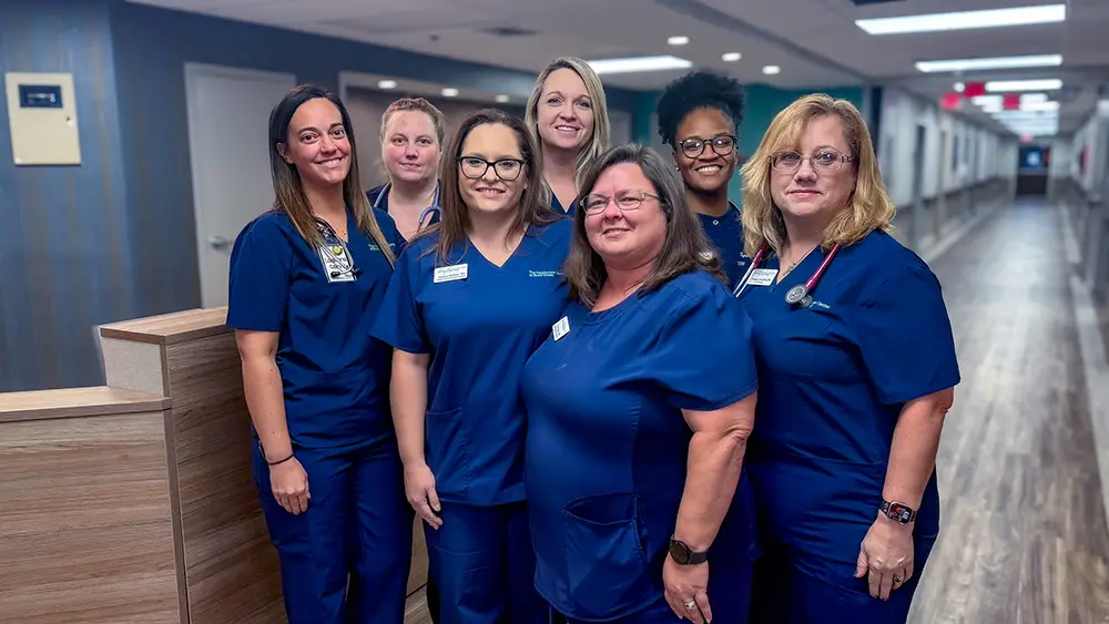 Seven healthcare workers in blue scrubs stand together and smile for a group photo in a hospital hallway.